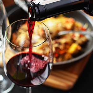 Un primer plano de 300x300 Pour Tinto vertido de una botella a una copa de vino. El fondo está ligeramente borroso y muestra un plato con comida sobre una tabla de madera. Los colores son ricos, con el rojo intenso del vino contrastando con el fondo oscuro y vibrante.