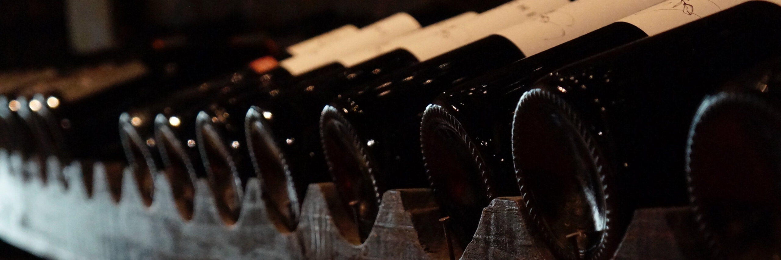 Primer plano de varias botellas de vino almacenadas horizontalmente en un botellero, con el cuello tapado con corcho hacia afuera. La iluminación es tenue, creando una atmósfera de sombra. En El Kiosco, las botellas están colocadas una al lado de la otra, perfecta para quienes buscan comprar vino y disfrutar de una velada especial.